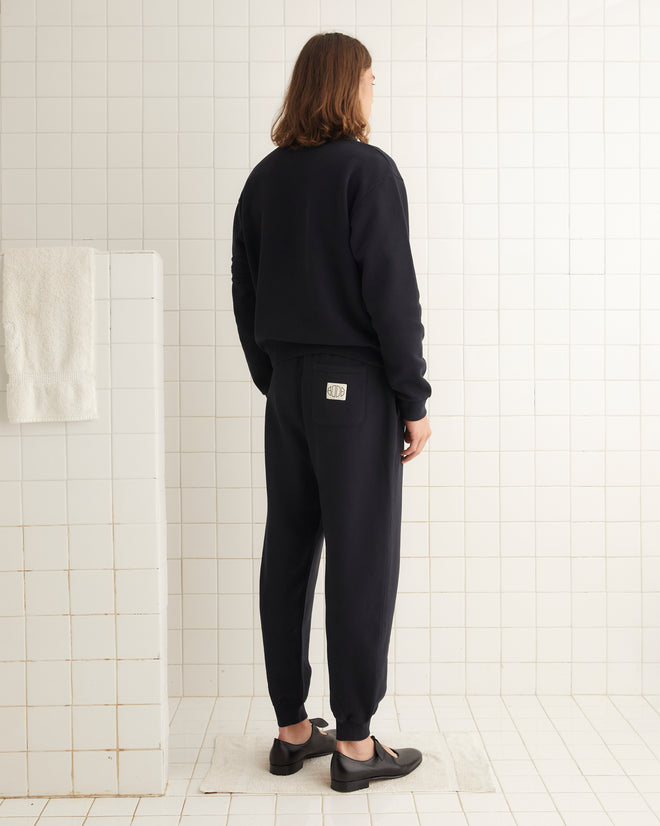 The person with long hair, dressed in a Crew Polo - Black and black jogger pants, stands facing forward in a tiled room, capturing the relaxed essence of 1980s rugby shirts.