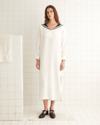 A person stands in a white tiled room, wearing the Cricket Dress—a long sailor dress featuring a navy collar—paired with black shoes, capturing the timeless charm of 1970s women's sportswear.