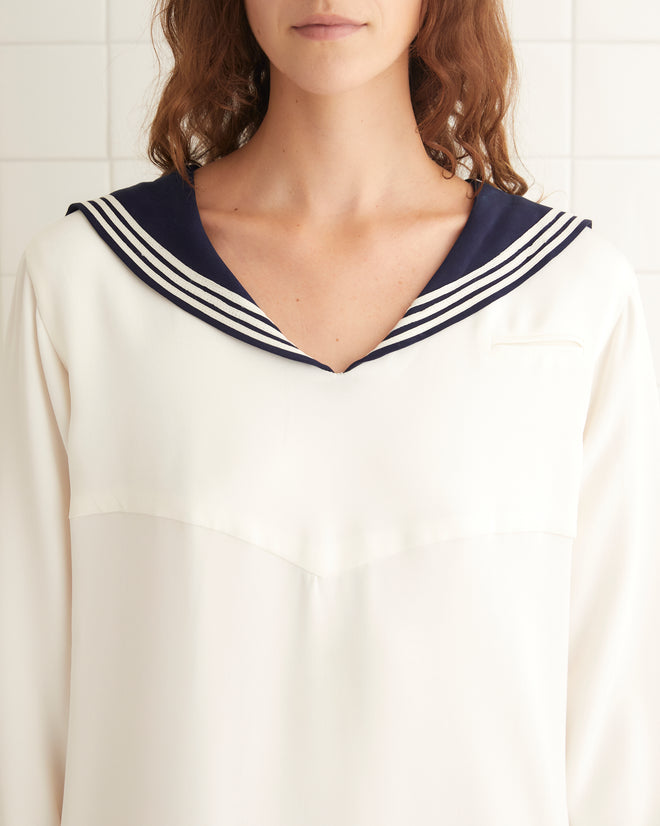 A person stands in a white tiled room, wearing the Cricket Dress—a long sailor dress featuring a navy collar—paired with black shoes, capturing the timeless charm of 1970s women's sportswear.