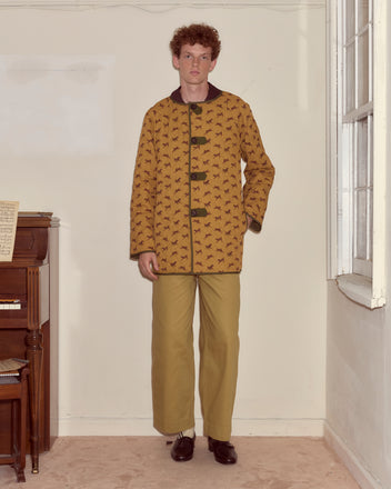 A man wearing the Crittendon Liner Coat and khaki pants stands by a piano in a beige room with a window.