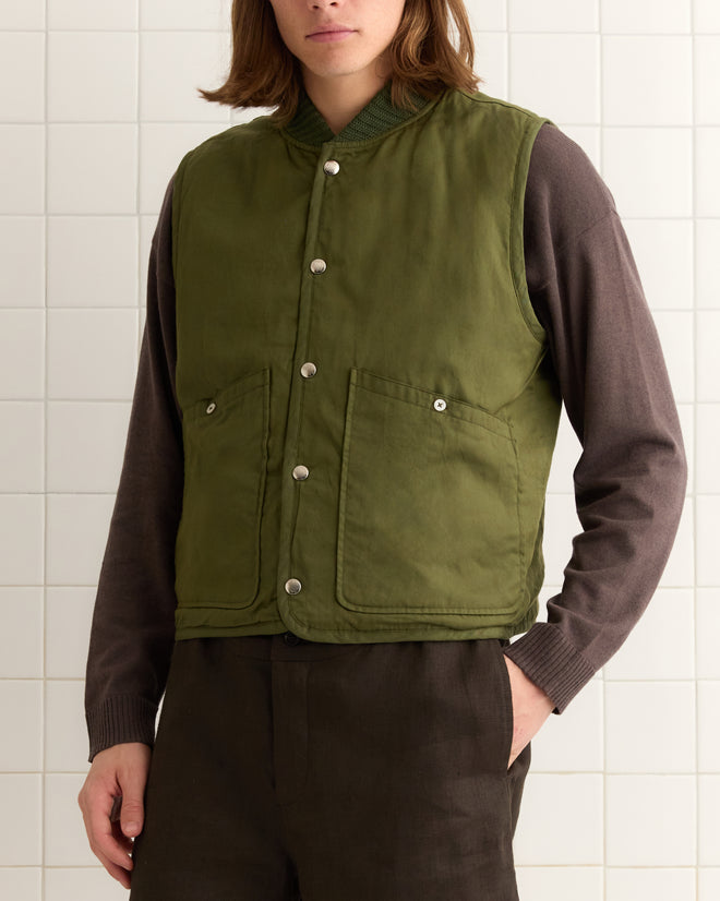 A person wearing a Crittendon Quilted green, reversible vest with Bode snap buttons over a brown long-sleeve shirt stands against a tiled wall.