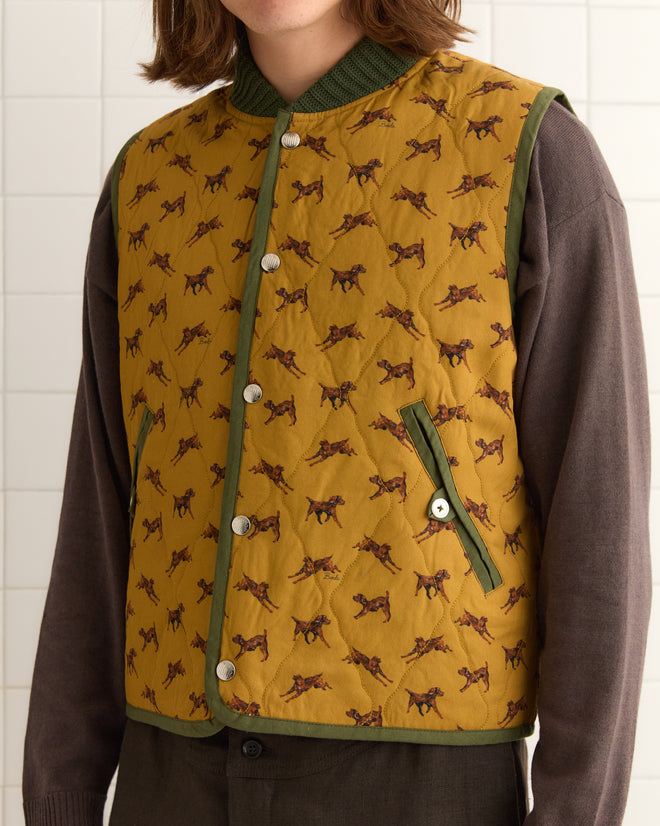 Wearing the Crittendon Quilted Vest adorned with horse patterns and Bode snap buttons, a person stands against a tiled wall, the vest layered over a brown long-sleeve shirt.