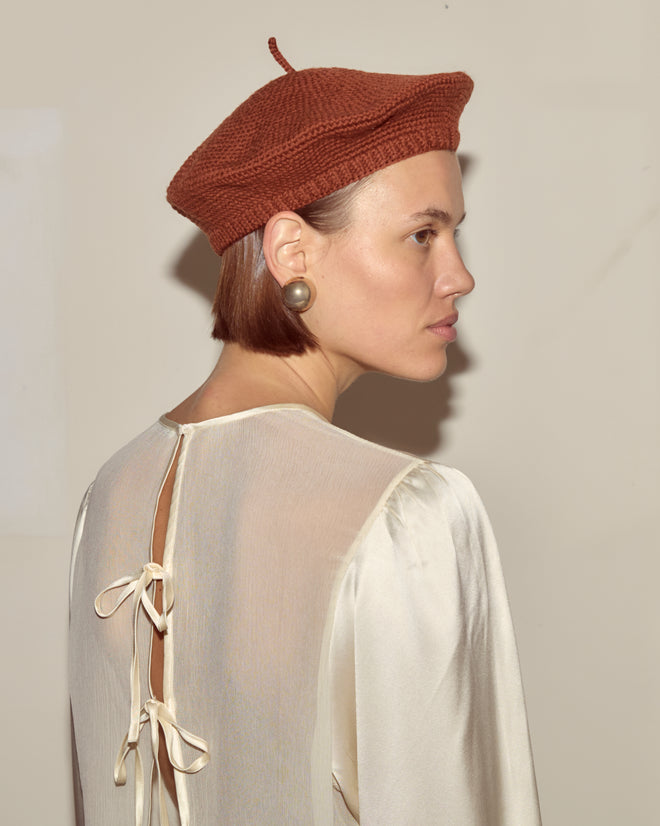 Woman wearing a Crochet Beret - Tobacco, hand-knit from merino wool, styled with a cream blouse featuring back bows, facing sideways against a plain background.