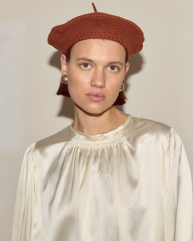 Woman in a cream satin blouse and pearl earrings, accessorized with the Crochet Beret - Tobacco, standing against a plain background.