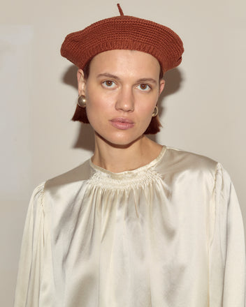 A woman poses against a plain background wearing a Crochet Beret in Tobacco, pearl earrings, and a cream satin blouse.