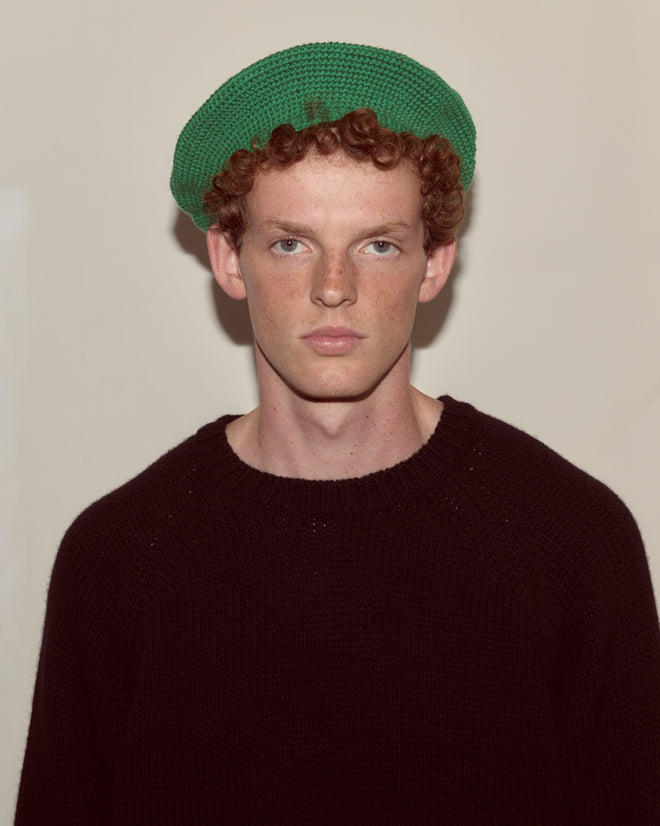 A young person with curly red hair, wearing a green Crochet Beret and a black sweater, looks at the camera.