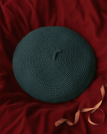 The Crochet Beret - Green sits stylishly on a red fabric background, adorned with a delicate peach ribbon.