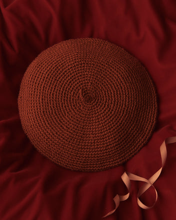 The Crochet Beret - Tobacco, a round and textured brown hat adorned with a ribbon, rests gracefully on the red fabric, exuding elegance and charm.