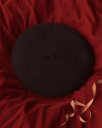 A black Crochet Beret made of merino wool rests on red fabric, with a curled pink ribbon beside it.