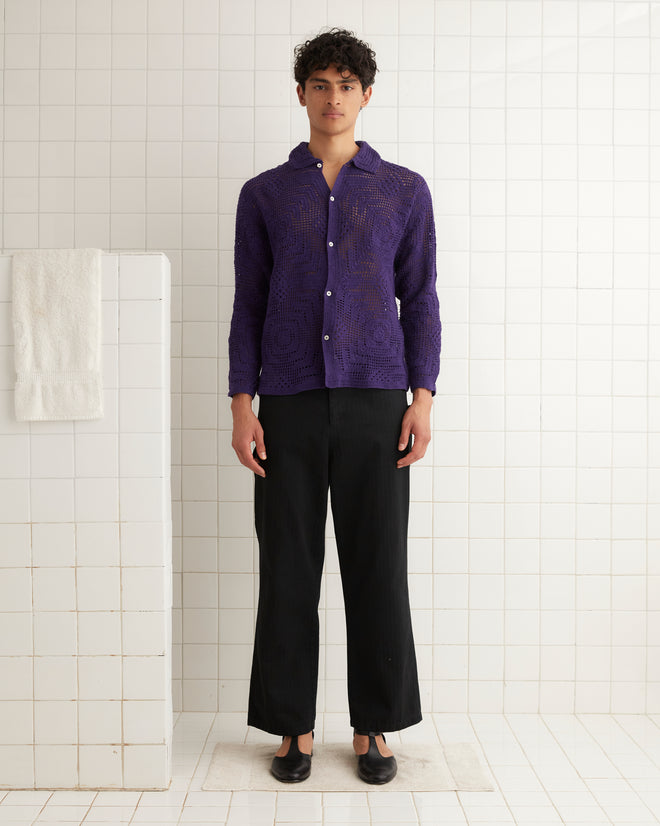 A person in a Crochet Overshirt - Purple and black pants stands in a white tiled room with a towel hanging nearby.