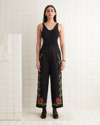 Person wearing a black top and Ribboned Hollyhock Trousers, featuring embroidered colorful side patterns, standing in a tiled room.