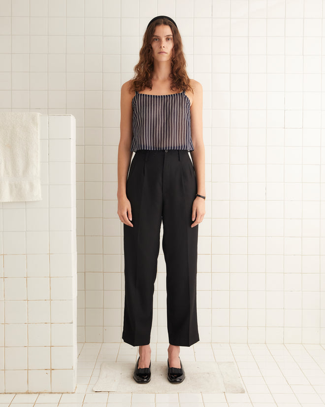 A woman with long wavy hair stands in a tiled room, wearing a Croquet Field Tank - S made of lightweight cotton voile, black high-waisted trousers, and black shoes. A white towel hangs on a partition beside her.