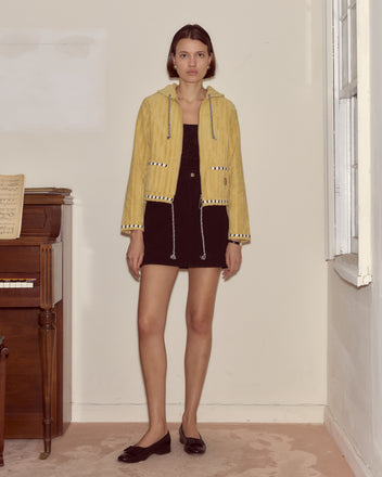 Woman in a yellow jacket and black dress stands indoors beside a piano and window, her look elevated by the Croquet Ground Hoodie.