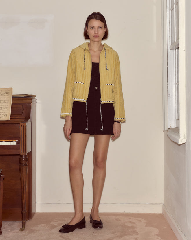 Woman in a yellow jacket and black dress stands indoors beside a piano and window, her look elevated by the Croquet Ground Hoodie.