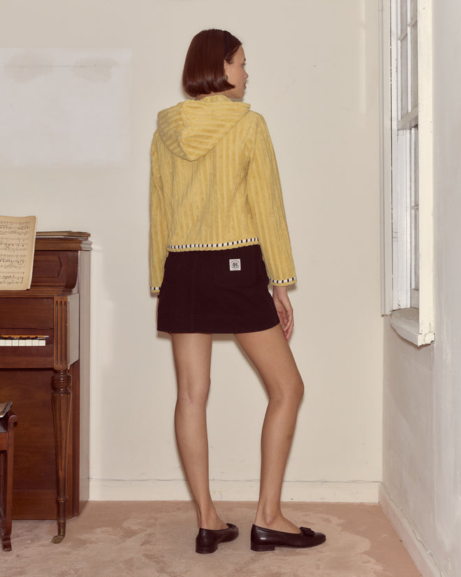 A woman stands indoors in a green Croquet Ground Hoodie with an embroidered Bode logo and black skirt, facing away near a piano and window.