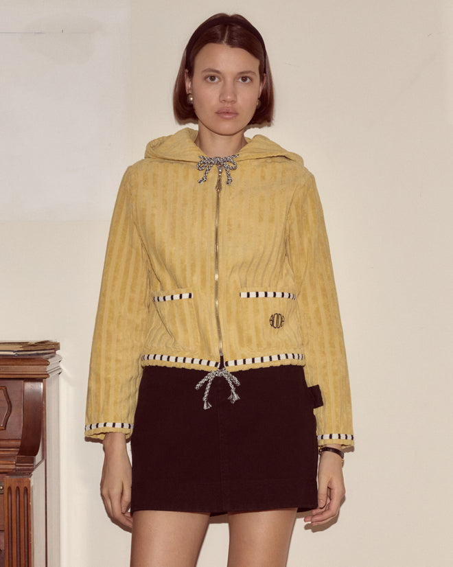 A woman in a yellow Croquet Ground Hoodie and black skirt stands indoors against a plain wall, looking at the camera.