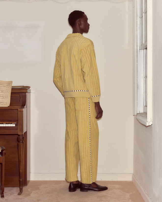 A person in the Croquet Ground Pullover, a yellow striped cotton sweatshirt with an embroidered Bode logo, stands facing a corner near a window and a piano with sheet music.