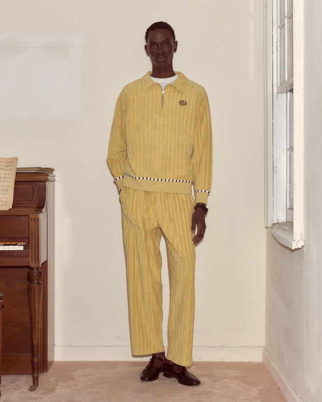 A person stands indoors near a piano and window, wearing a yellow striped tracksuit, brown shoes, and the Croquet Ground Pullover—a cotton sweatshirt with an embroidered Bode logo.