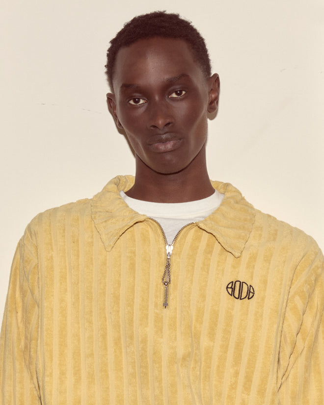 A person wears the Croquet Ground Pullover— a yellow cotton sweatshirt with an embroidered Bode logo— standing against a plain off-white background.