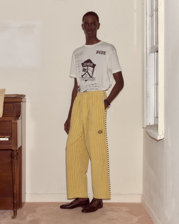A person stands indoors by a piano wearing Croquet Ground Sweatpants—loose green terrycloth pants with an embroidered Bode logo—and a graphic white T-shirt.