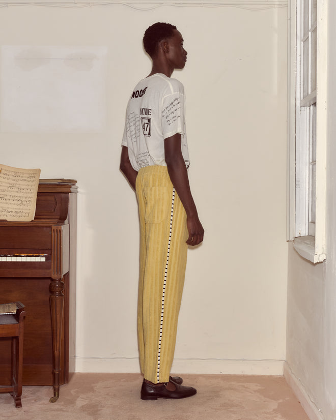 A man in a graphic tee and Croquet Ground Sweatpants stands sideways by a piano near a window in a simple room.