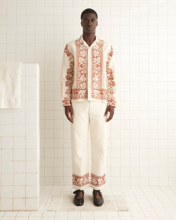 A man stands in a tiled room, donning an elegant cream-colored outfit made of 100% linen. The ensemble, comprising the Cross-Stitched Rose Garland Long Sleeve Shirt and matching pants, is adorned with intricate red cross-stitching and features an embroidered rose border that adds a refined touch to his appearance.