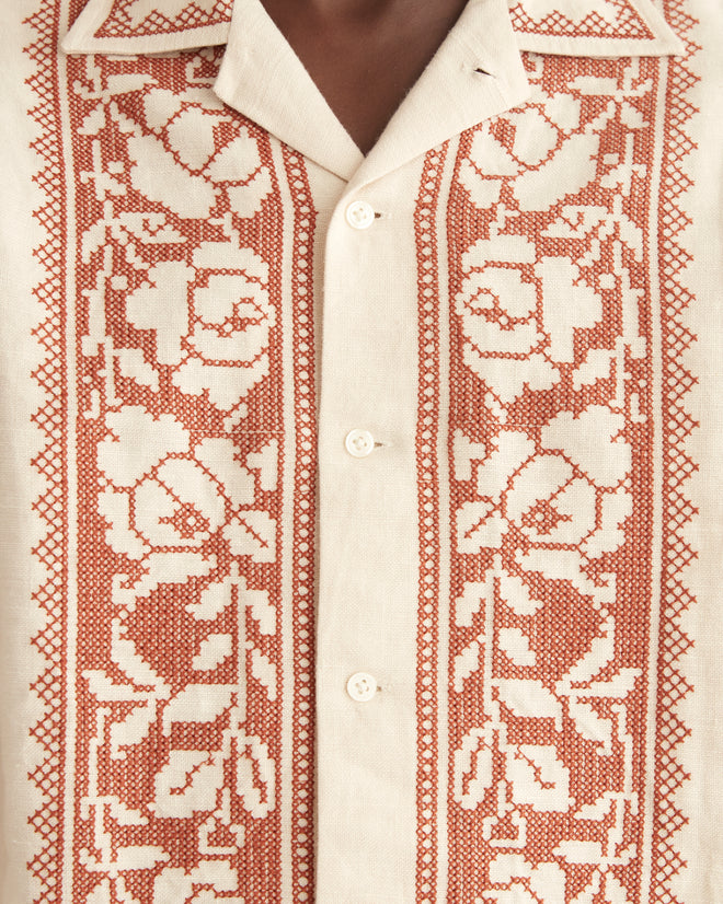 A man stands in a tiled room, donning an elegant cream-colored outfit made of 100% linen. The ensemble, comprising the Cross-Stitched Rose Garland Long Sleeve Shirt and matching pants, is adorned with intricate red cross-stitching and features an embroidered rose border that adds a refined touch to his appearance.