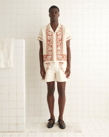 A person stands in a white tiled room, wearing a Cross-Stitched Rose Garland Short Sleeve Shirt made from 100% linen, featuring intricate brown floral patterns. The coordinated outfit includes matching shorts, creating a sophisticated and timeless look with its vintage-inspired design.