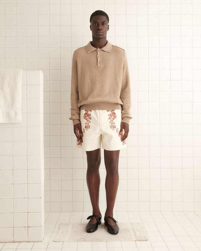 A man stands in a white-tiled room wearing a beige long-sleeve polo shirt, Cross-Stitched Rose Garland Shorts, and dark sandals. His attire echoes a traditional rugby silhouette. He stands on a white bath mat near a tiled structure with an embroidered rose border and a folded 100% cotton towel.