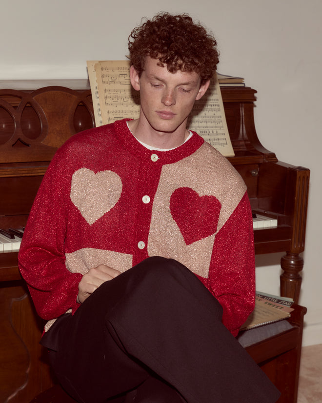A person with curly hair sits by a piano, wearing the Lamé Hearts Cardigan—a red lightweight sweater adorned with large heart shapes, evoking an Alice in Wonderland vibe.
