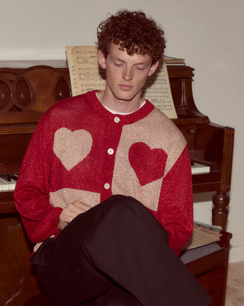 A man in a Lamé Hearts Cardigan sits by a piano with sheet music, capturing an Alice in Wonderland-inspired whimsical charm.