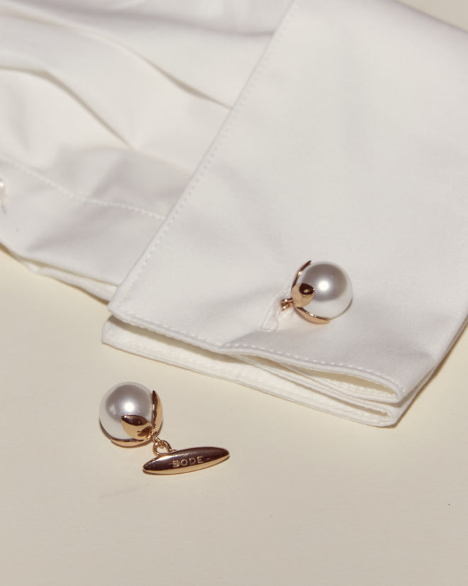 A white shirt cuff with Curio Cufflinks - White, one matching cufflink nearby featuring a gold tag engraved 