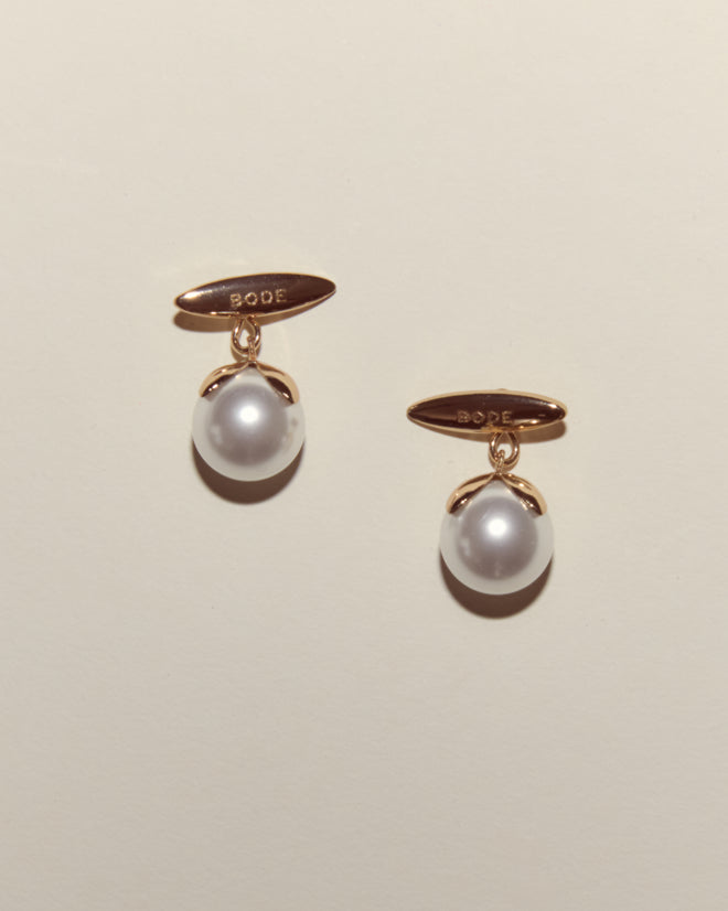 Curio Cufflinks - White: Gold earrings featuring engraved tags and single white pearls, inspired by Victorian Revival cufflinks, showcased on a light beige background.