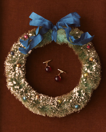 A festive wreath decorated with a blue bow and colorful ornaments features two Curio Cufflinks in red at the center, set against a brown background.