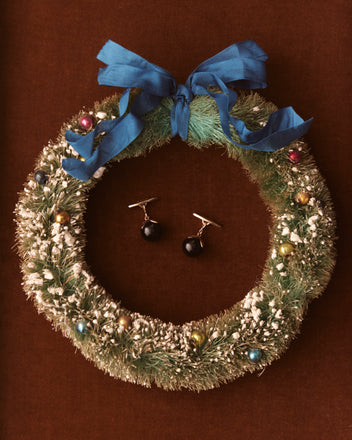 A festive wreath adorned with a blue bow and colorful ornaments is displayed against a brown background alongside the Curio Cufflinks - Black.