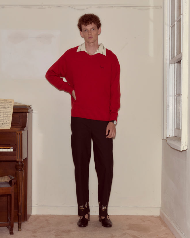 A man wearing the Currant Sweater - Red and black pants stands indoors by a piano and a window.
