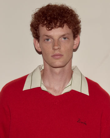 A young man with curly red hair wears the Currant Sweater - Red, a wool cashmere blend V-neck, over a collared shirt, standing against a plain background.