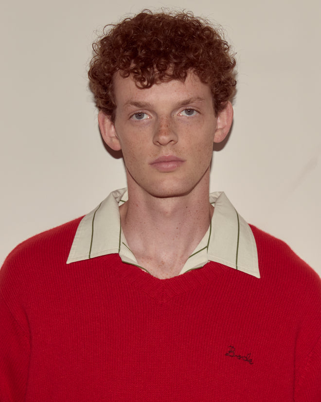 A young man with curly red hair wears the Currant Sweater - Red, a wool cashmere blend V-neck, over a collared shirt, standing against a plain background.