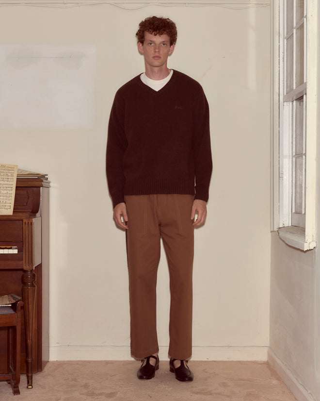 A man stands indoors next to a piano and window wearing the Currant Sweater – Dark Brown, brown pants, and black shoes.