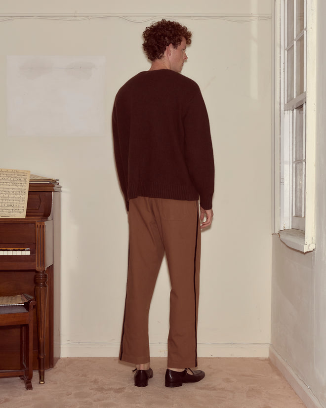 A man stands indoors facing away, wearing the Currant Sweater in dark brown and tan pants next to an upright piano.