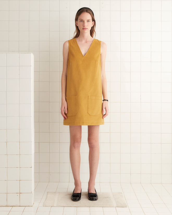 The Custom Senior Cord Dress in mustard hangs elegantly on a hanger against a backdrop of white tiled walls, reminiscent of the classic British corduroy style.