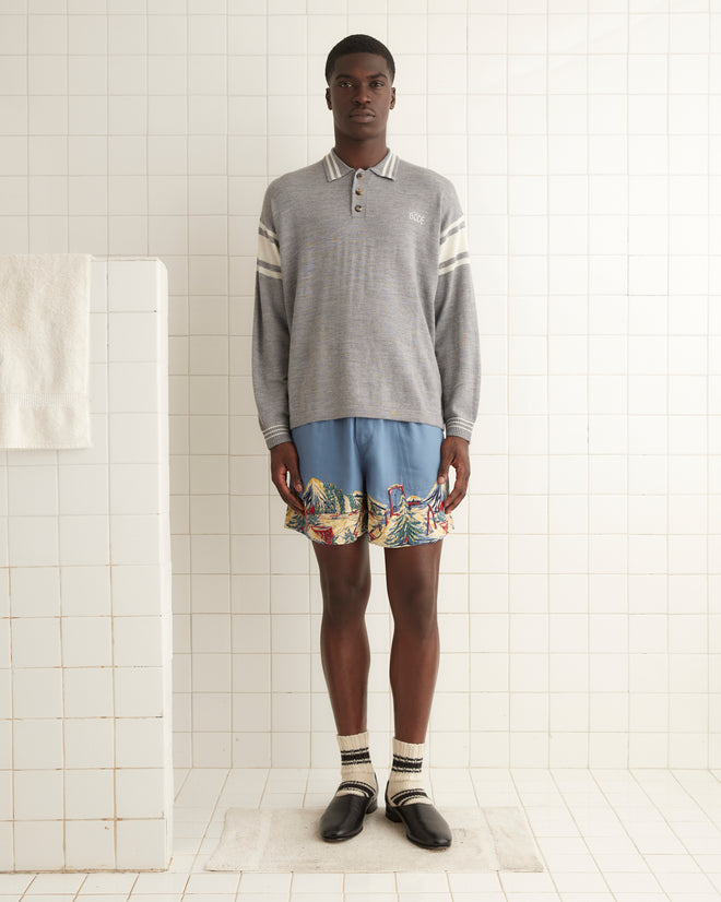 A person stands in a tiled room wearing a gray long-sleeve Cycling Polo, blue shorts with a colorful print resembling a vintage cycling jersey, ankle socks, and black loafers. A white towel hangs on a rack to the left.