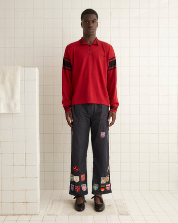 A person stands in front of a tiled wall, wearing a red Cycling Polo - Red made of merino wool with long sleeves and black pants adorned with multiple patches.