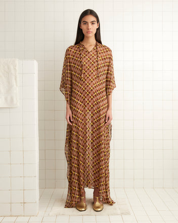 A woman in a Cattail Plaid Kaftan and gold shoes stands in a tiled room with a towel on the wall.