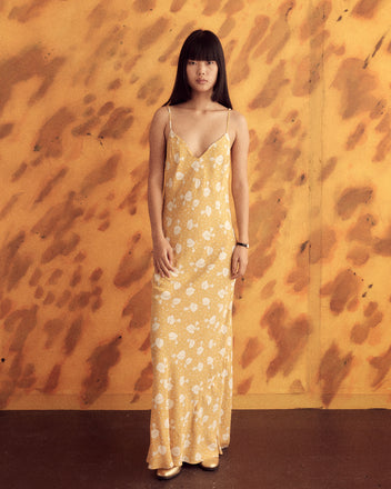 A woman in a Cinquefoil Cluster Slip - 8, featuring golden yellow spaghetti straps and a white floral pattern reminiscent of vintage 1940s fabric, stands against a matching yellow and brown abstract background.