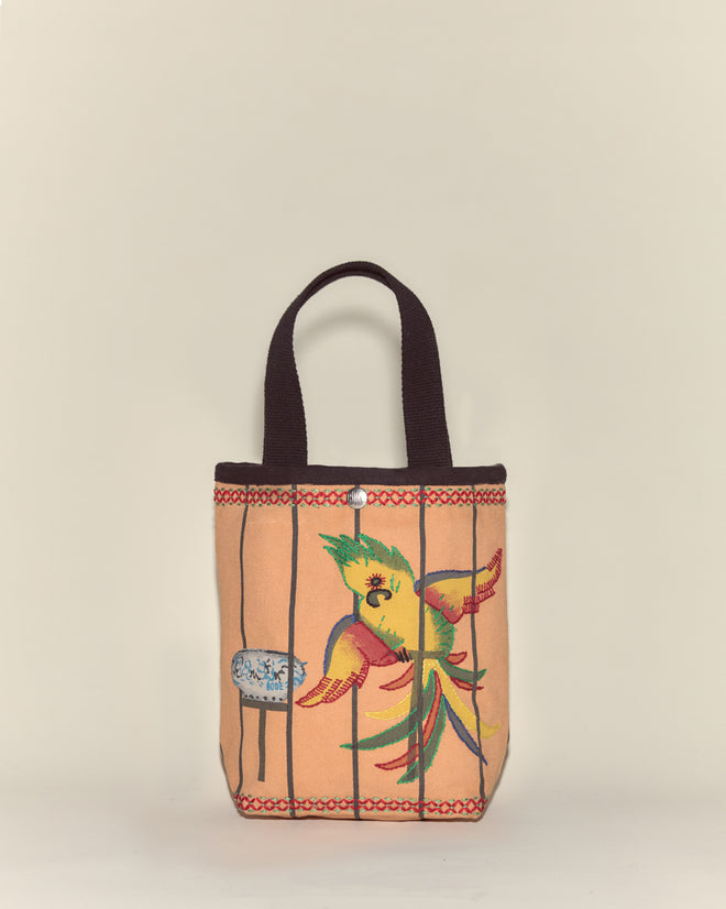The Cockatoo Small Tote is a canvas bag with black handles, featuring a vibrant painted cockatoo and water dish inside a cage design.