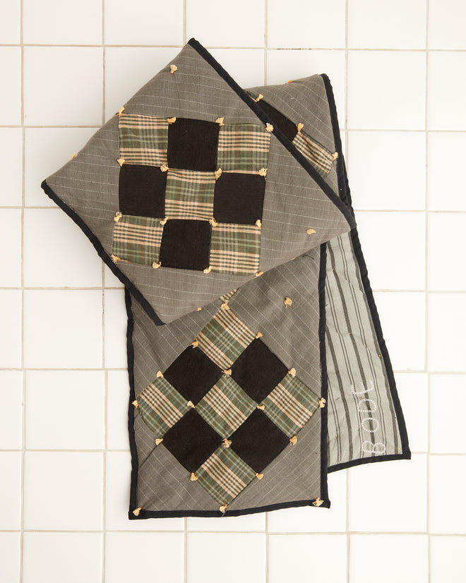 A pair of Corduroy Nine Patch Quilt Scarf pillows, with a black and green plaid design inspired by 1920s-1930s quilting, rest elegantly on a white tiled floor.