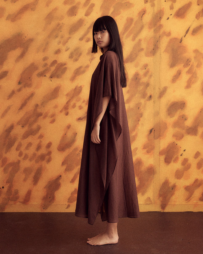 A person with long dark hair stands barefoot wearing the Cove Kaftan, which has a 1970s-inspired design, against a yellow and brown textured background.