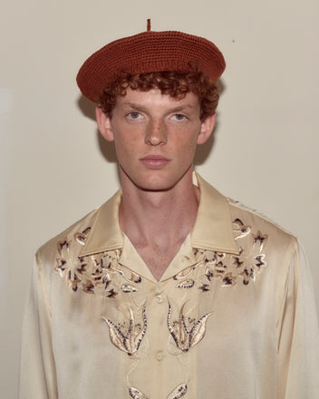 A person wearing a Crochet Beret - Tobacco and a cream embroidered shirt stands against a plain beige background.