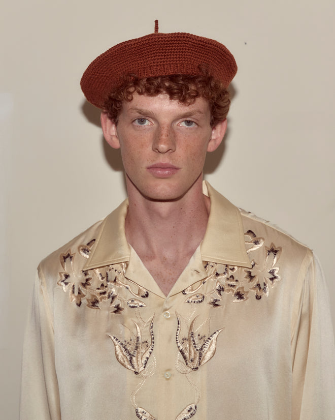 A person wearing a Crochet Beret - Tobacco and a cream embroidered shirt stands against a plain beige background.